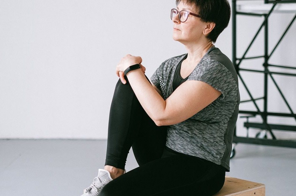 Senior woman in active wear sitting on a wooden box indoors, reflecting on wellness.